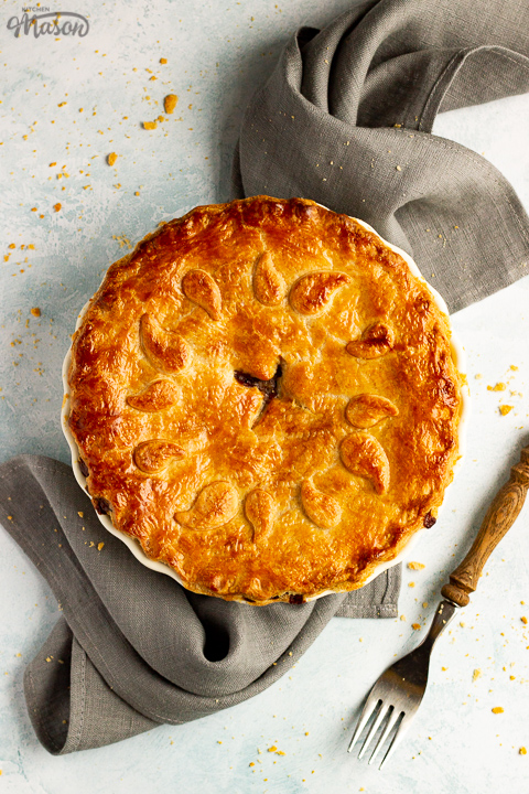 A baked vegetarian mince and onion pie decorated with pastry leaves sat on a grey linen napkin with a wooden handled fork. Set on a pale blue wash backdrop.