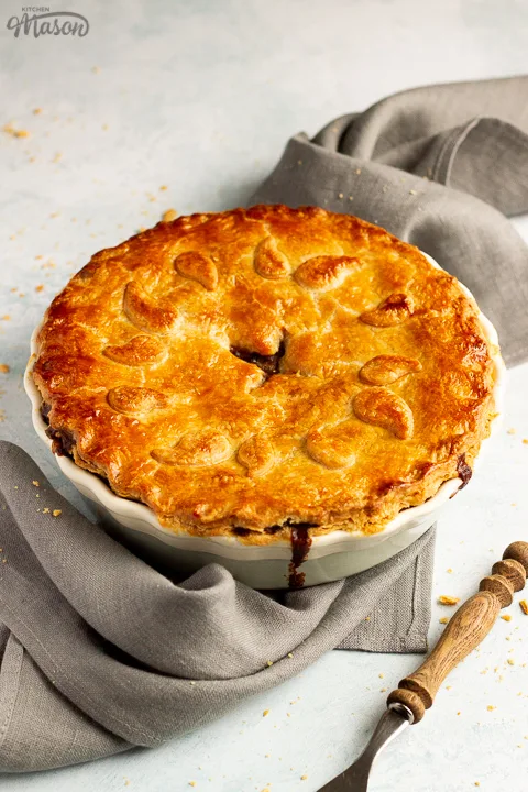 A baked vegetarian mince and onion pie decorated with pastry leaves sat on a grey linen napkin with a wooden handled fork. Set on a pale blue wash backdrop.