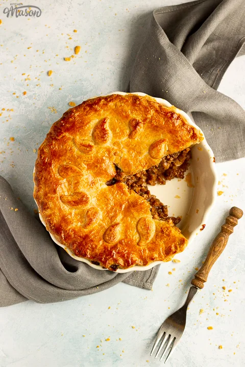 A baked vegetarian mince and onion pie with a slice cut out, decorated with pastry leaves, sat on a grey linen napkin with a wooden handled fork. Set on a pale blue wash backdrop.