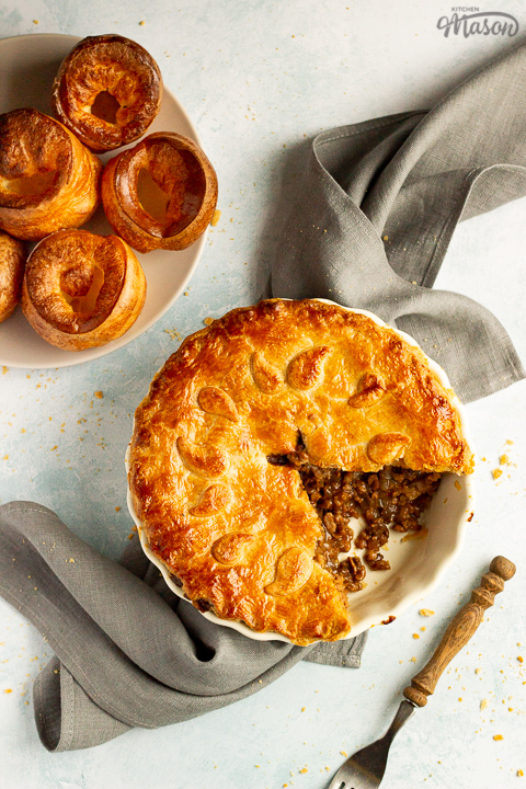 A baked vegetarian mince and onion pie with a slice cut out, decorated with pastry leaves, sat on a grey linen napkin. There's a wooden handled fork and a plate of Yorkshire puddings in the background. Set on a pale blue wash backdrop.
