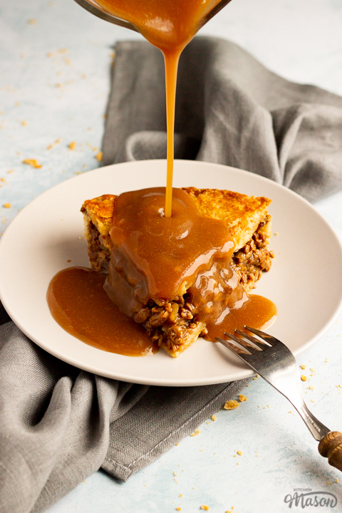 A slice of vegetarian mince and onion pie on a light grey plate with a wooden handled fork resting on it and gravy being poured onto it. Set on a grey linen napkin over a pale blue wash backdrop.