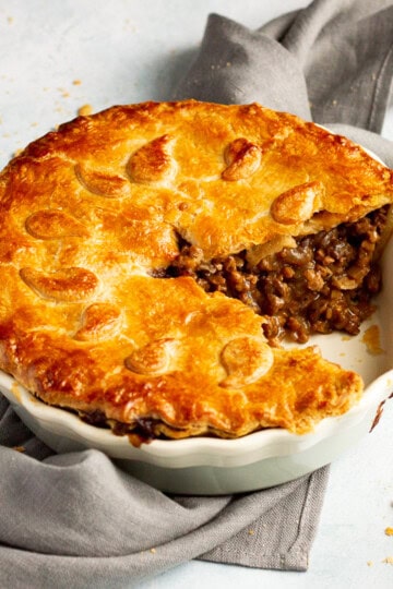 A baked vegetarian mince and onion pie with a slice cut out, decorated with pastry leaves, sat on a grey linen napkin with a wooden handled fork. Set on a pale blue wash backdrop.