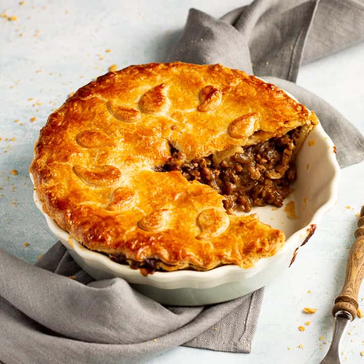 A baked vegetarian mince and onion pie with a slice cut out, decorated with pastry leaves, sat on a grey linen napkin with a wooden handled fork. Set on a pale blue wash backdrop.