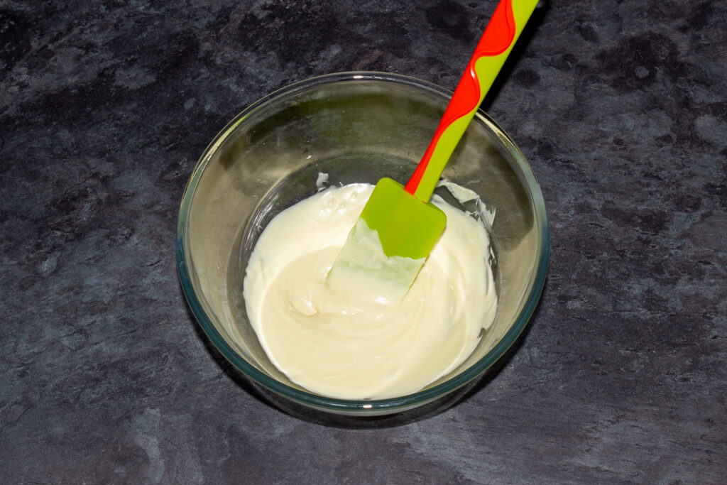 Melted white chocolate in a glass bowl with a green spatula.