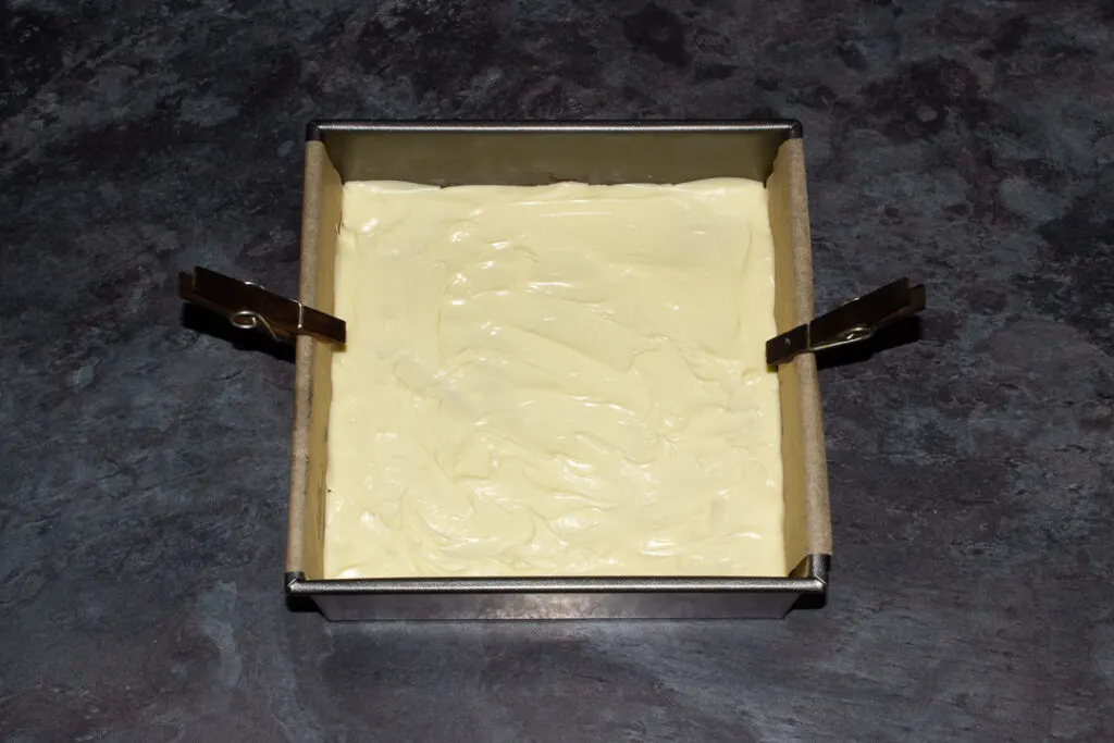 A lined square baking tin with melted white chocolate poured over caramel and red velvet brownie.