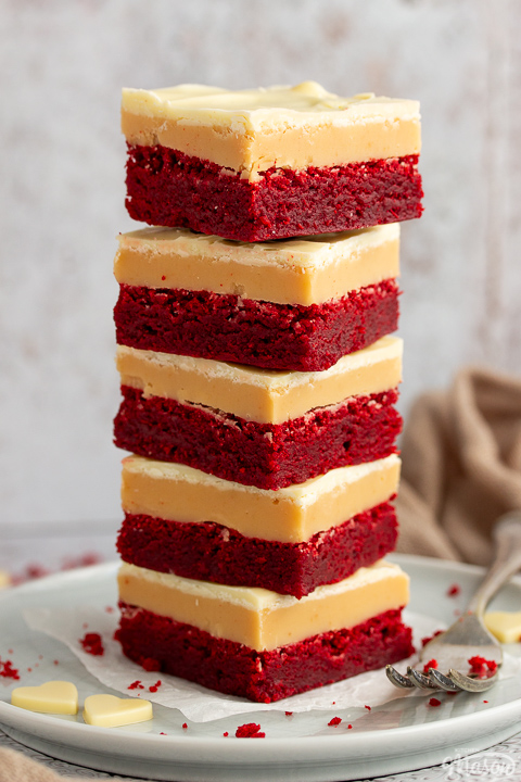 A stack of 5 red velvet millionaire brownies set on two stacked plates with a fork on the side. There are white chocolate hearts, red velvet brownie crumbs and a light brown napkin in the background.