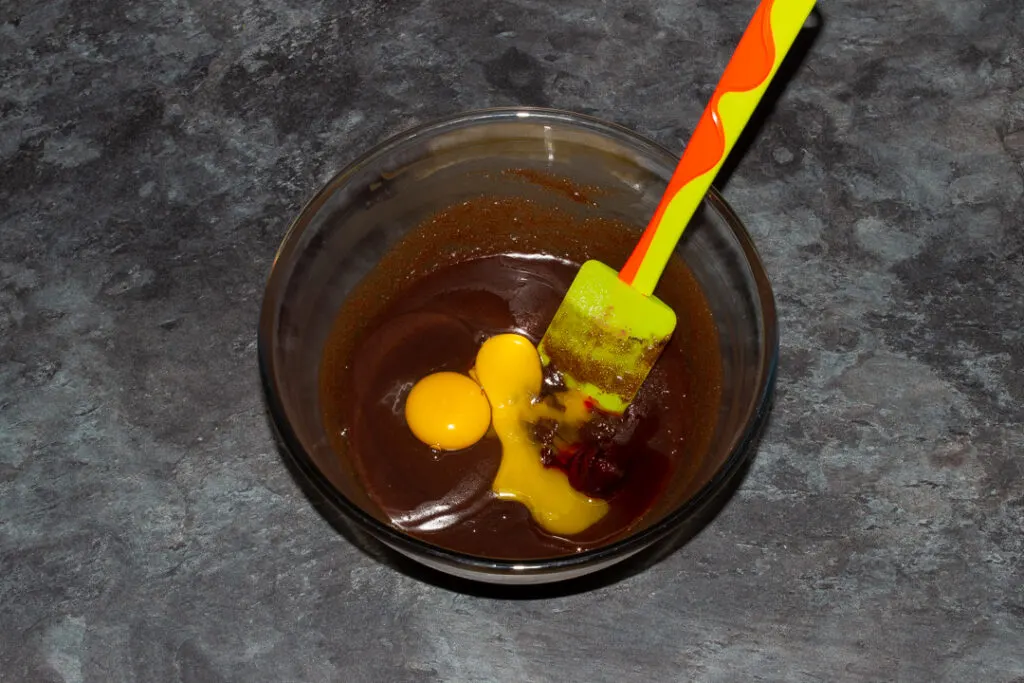 Egg, egg yolk, vanilla and red food colouring gel on top of a melted butter, sugar and cocoa powder mixture in a glass bowl with a green spatula.