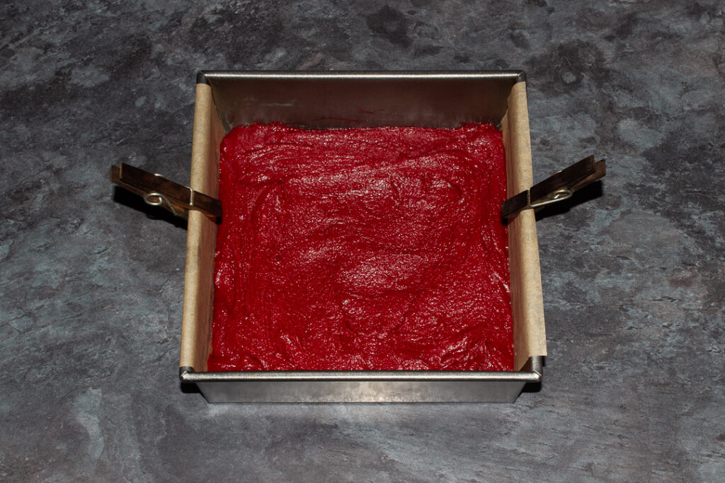 Red velvet brownie batter in a lined square baking tin with two pegs on each side.