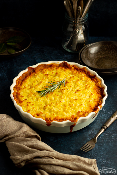 Vegetarian shepherd's pie in a dish topped with a sprig of rosemary. There's cutlery, dark bowls and a light brown napkin set over a deep blue backdrop.