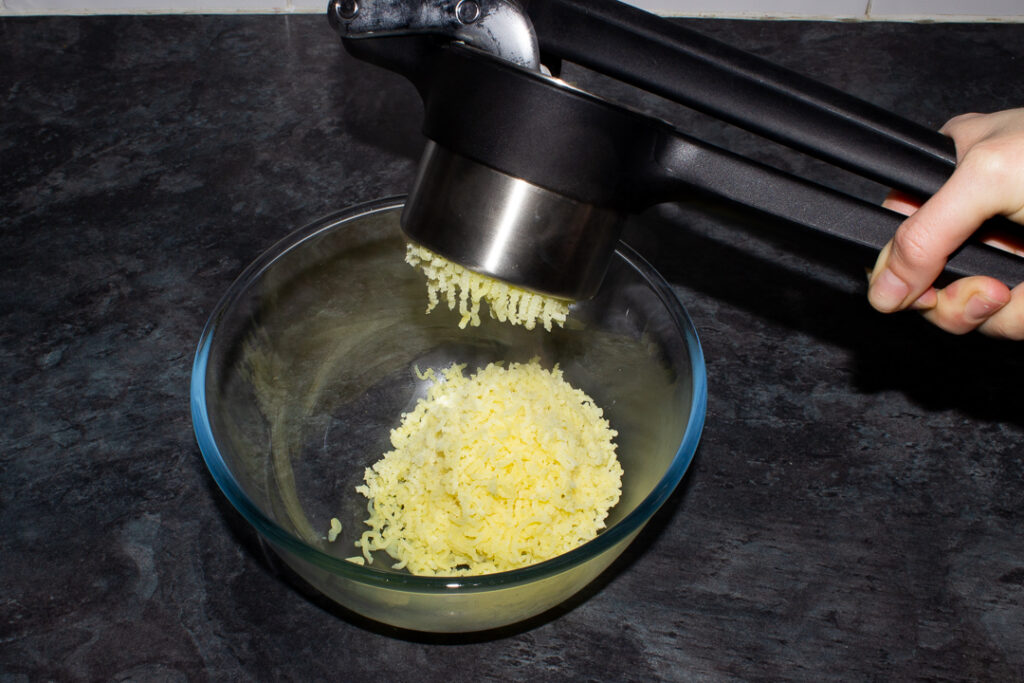Boiled potatoes being pressed through a potato ricer into a glass bowl on a kitchen worktop