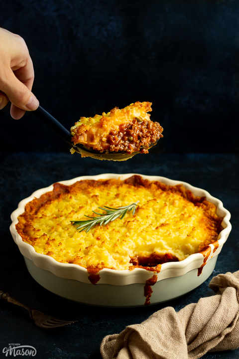 Vegetarian shepherd's pie on a deep blue backdrop with a light brown napkin. Someone is holding a large spoon full of the pie above it.