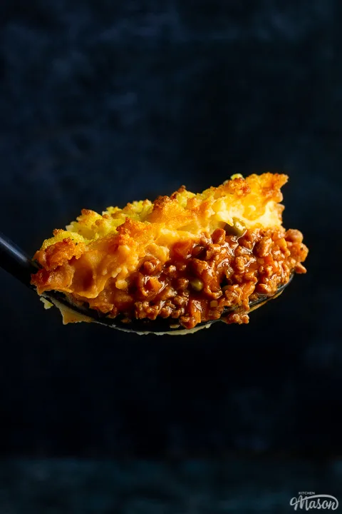 A close up of a large spoonful of vegetarian shepherd's pie up against a deep blue backdrop