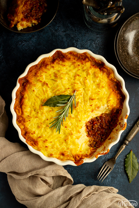 A birds eye view of vegetarian shepherd's pie with a spoonful taken out of it. There are dark bowls, cutlery bay leaves and a light brown napkin set over a deep blue backdrop.