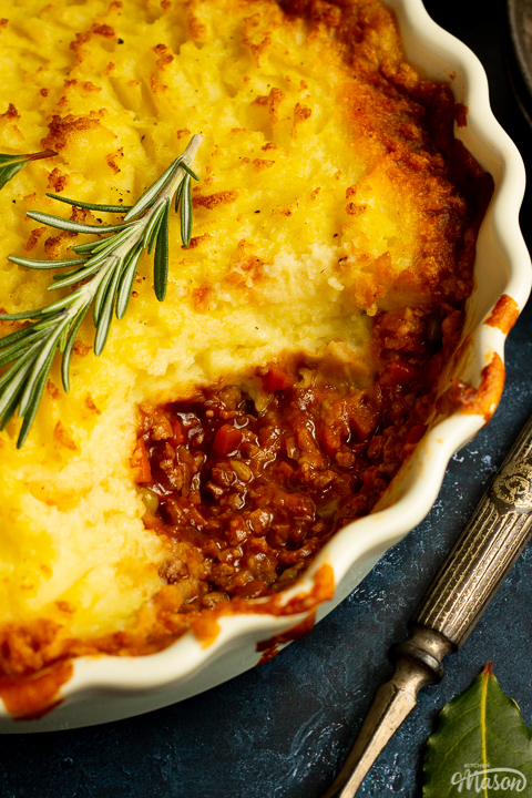 A close up view of vegetarian shepherd's pie topped with a rosemary sprig with a spoonful taken out of it. There's a fork and a dark bowl in the background set over a deep blue backdrop,