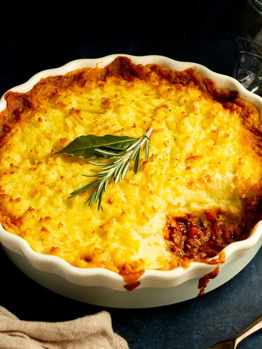 Vegetarian shepherd's pie in a dish with a spoonful taken out of it and a sprig of rosemary on top. There's cutlery, dark bowls and a light brown napkin set over a deep blue backdrop.