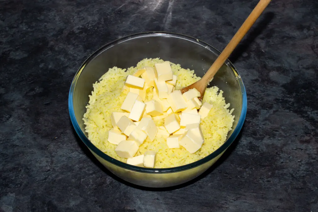 Boiled potato that's been pressed through a potato ricer and cubed butter in a glass bowl on a kitchen worktop