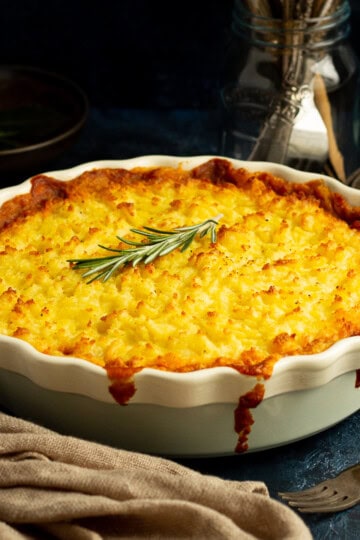 Vegetarian shepherd's pie in a dish topped with a sprig of rosemary. There's cutlery, dark bowls and a light brown napkin set over a deep blue backdrop.