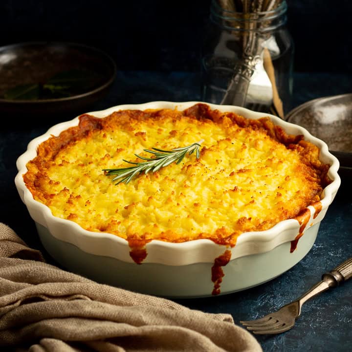Vegetarian shepherd's pie in a dish topped with a sprig of rosemary. There's cutlery, dark bowls and a light brown napkin set over a deep blue backdrop.