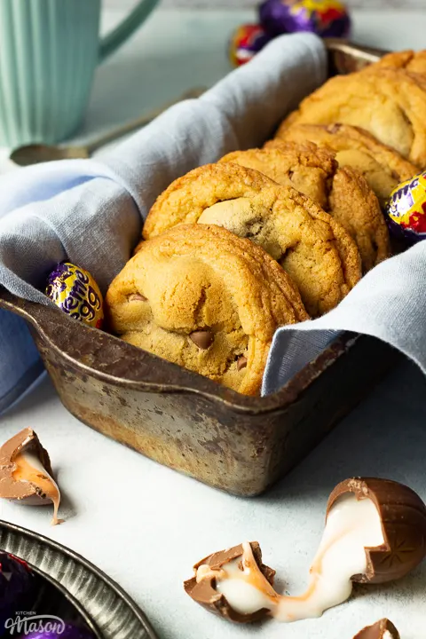 Creme Egg cooies in a bread tin lined with a pale blue napkin surrounded by whole and broken Creme Eggs. Set on a pale blue backdrop with a tray of whole Creme Eggs and a mug of tea in the background.