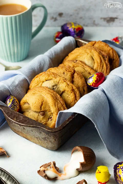 Creme Egg cookies in a bread tin lined with a pale blue napkin surrounded by whole and broken Creme Eggs. Set on a pale blue backdrop with a tray of whole Creme Eggs, a mug of tea and a yellow Easter chick in the background.