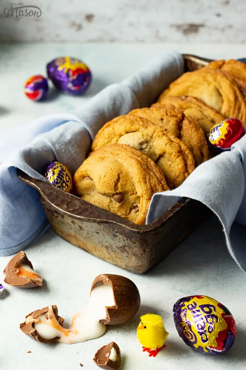 Creme Egg cookies in a bread tin lined with a pale blue napkin surrounded by whole and broken Creme Eggs. Set on a pale blue backdrop with a yellow Easter chick in the background.