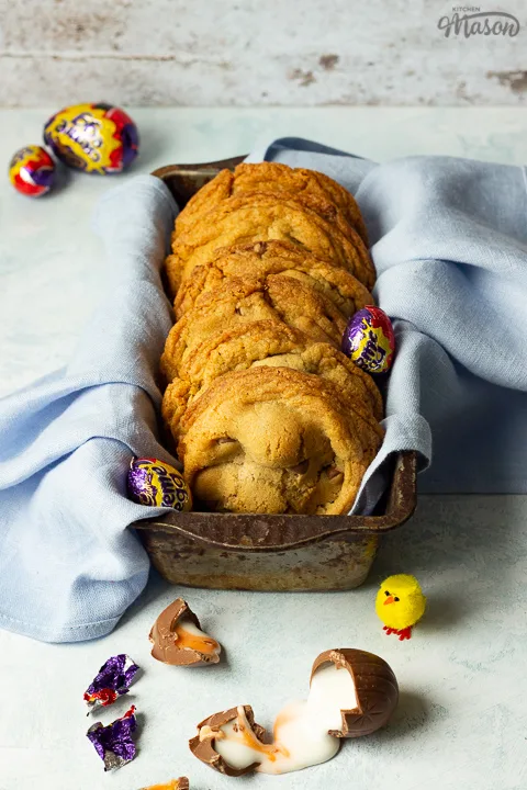 Front view of Creme Egg cookies in a bread tin lined with a pale blue napkin surrounded by whole and broken Creme Eggs. Set on a pale blue backdrop with a yellow Easter chick in the background.