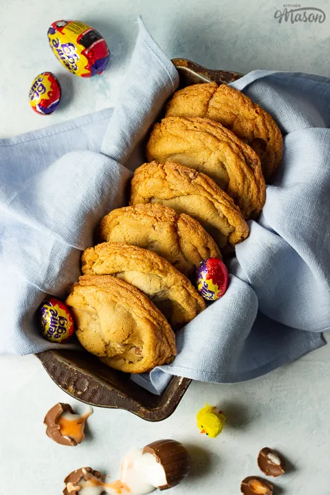 Birds eye view of Creme Egg cookies in a bread tin lined with a pale blue napkin surrounded by whole and broken Creme Eggs. Set on a pale blue backdrop with a yellow Easter chick in the background.