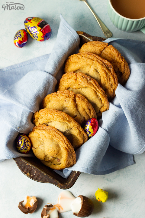 Birds eye view of Creme Egg cookies in a bread tin lined with a pale blue napkin surrounded by whole and broken Creme Eggs. Set on a pale blue backdrop with a mug of tea and a yellow Easter chick in the background.
