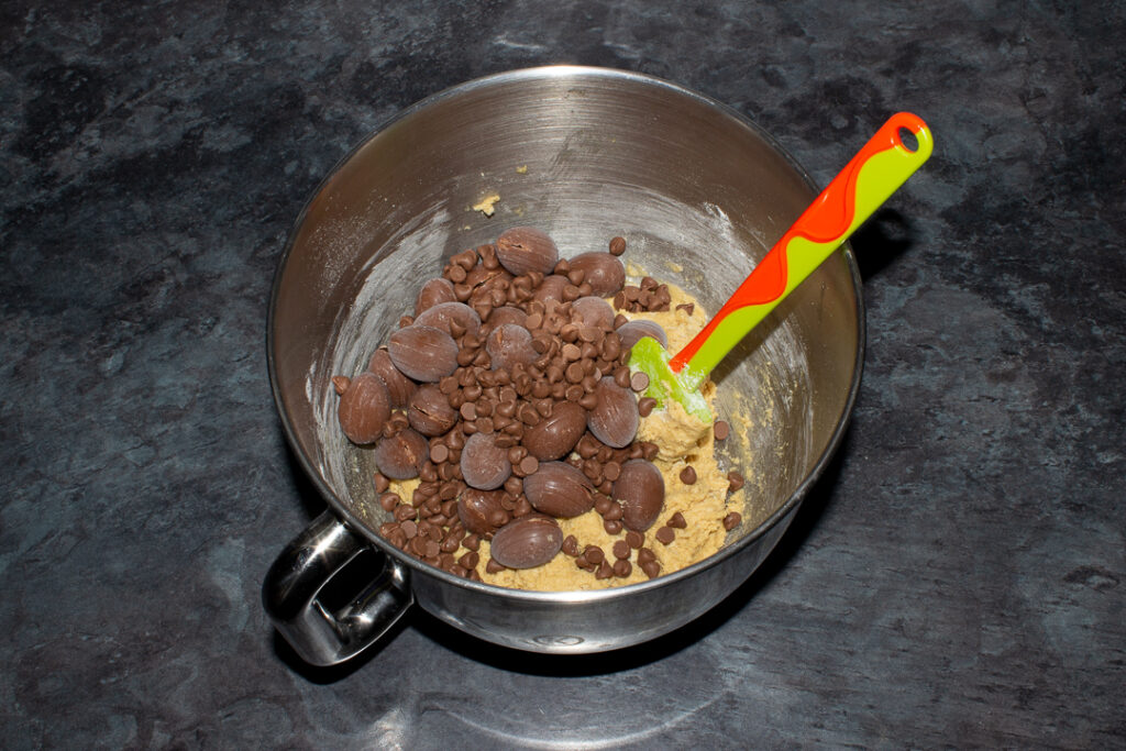 Frozen Creme Eggs and chocolate chips being added to cookie dough in the bowl of an electric stand mixer with a green spatula