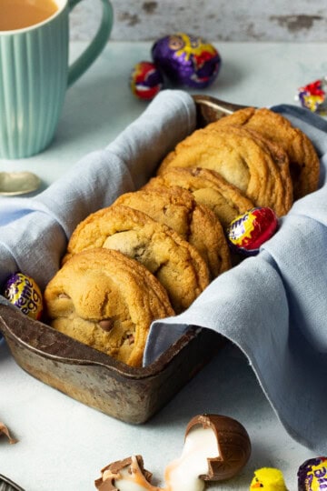 Creme Egg cookies in a bread tin lined with a pale blue napkin surrounded by whole and broken Creme Eggs. Set on a pale blue backdrop with a tray of whole Creme Eggs, a mug of tea and a yellow Easter chick in the background.