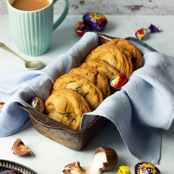 Creme Egg cookies in a bread tin lined with a pale blue napkin surrounded by whole and broken Creme Eggs. Set on a pale blue backdrop with a tray of whole Creme Eggs, a mug of tea and a yellow Easter chick in the background.