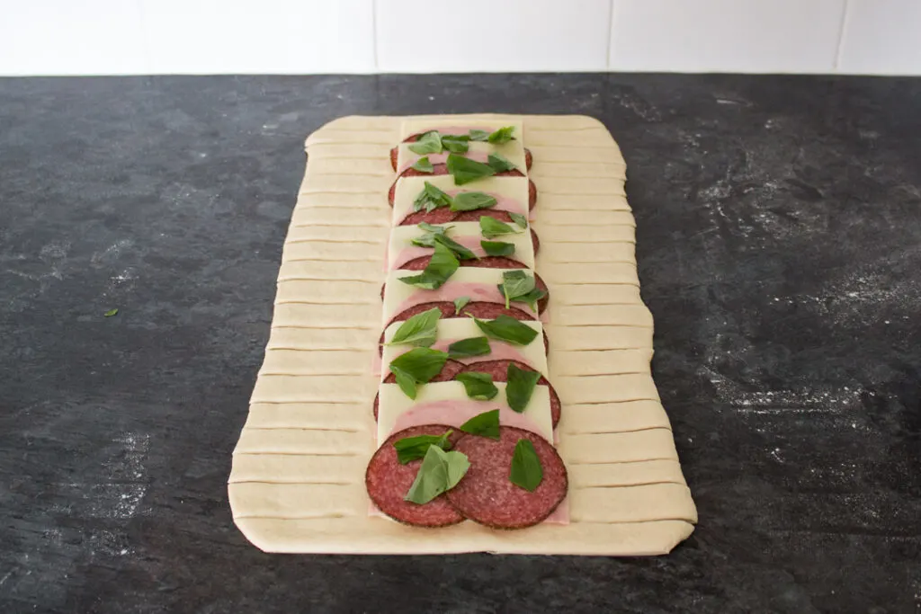 Pizza dough rolled out to a rectangle, topped with layered mozzarella, salami, ham and basil leaves on a lightly floured work surface. The dough has slices either side of the filling.
