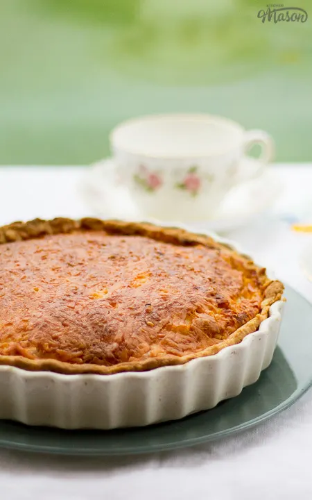 Gran's ham and cheese quiche in a white baking dish on a green plate. Set on a white floral table cloth with teacups and a saucer in the background.