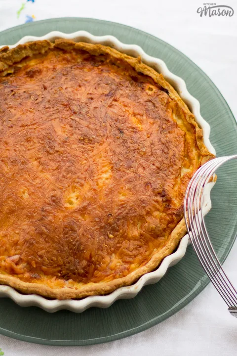 Gran's ham and cheese quiche in a white baking dish on a green plate. Set on a white floral table cloth with a pastry blender in the background.