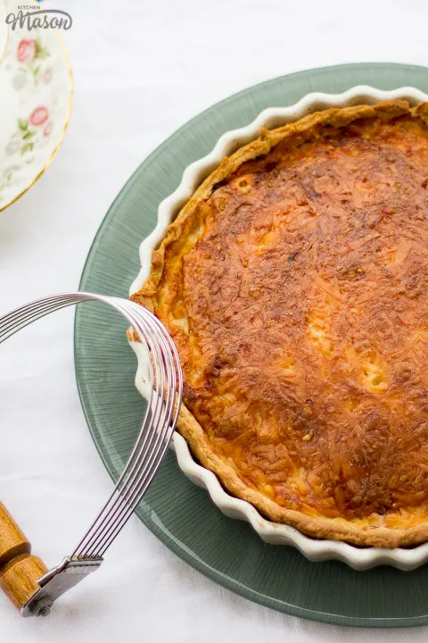 Gran's ham and cheese quiche in a white baking dish on a green plate. Set on a white floral table cloth with a pastry blender, teacups and a saucer in the background.