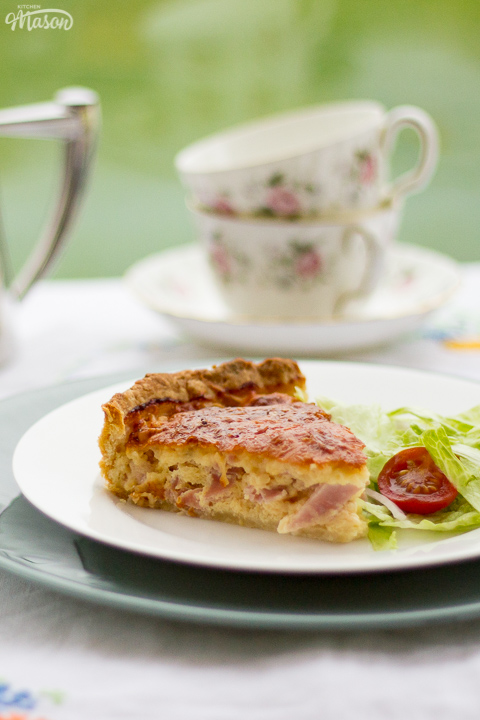 A slice of Gran's ham and cheese quiche and a side salad on a white plate set over a larger green plate. Set on a white floral table cloth with a teapot, cups and saucers in the background.
