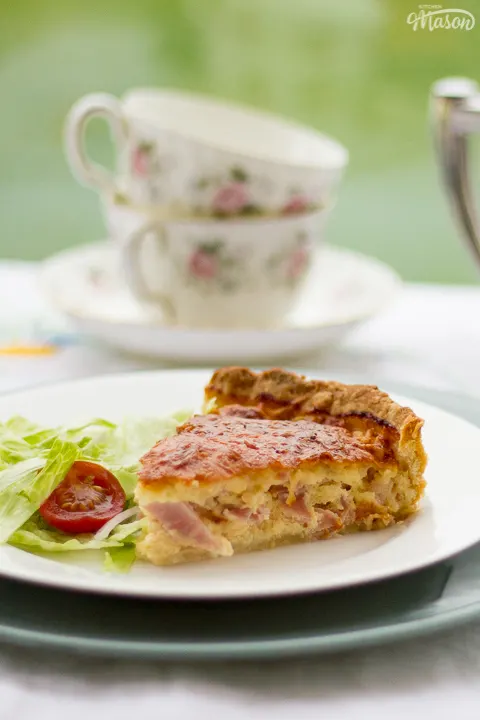 A slice of Gran's ham and cheese quiche and a side salad on a white plate set over a larger green plate. Set on a white floral table cloth with a teapot, cups and saucers in the background.