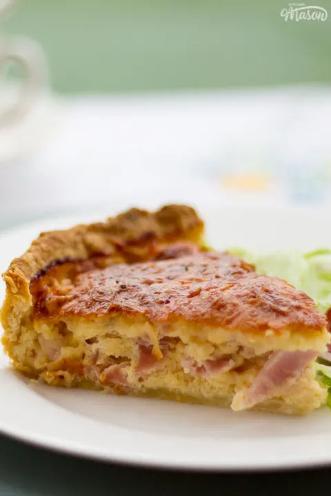 A slice of Gran's ham and cheese quiche and a side salad on a white plate set over a larger green plate. Set on a white floral table cloth with cups and saucers in the background.