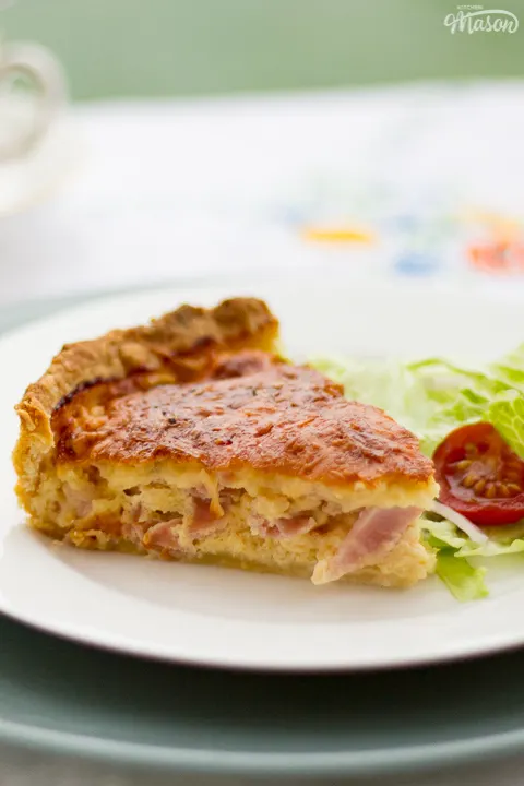 A slice of Gran's ham and cheese quiche and a side salad on a white plate set over a larger green plate. Set on a white floral table cloth with cups and saucers in the background.