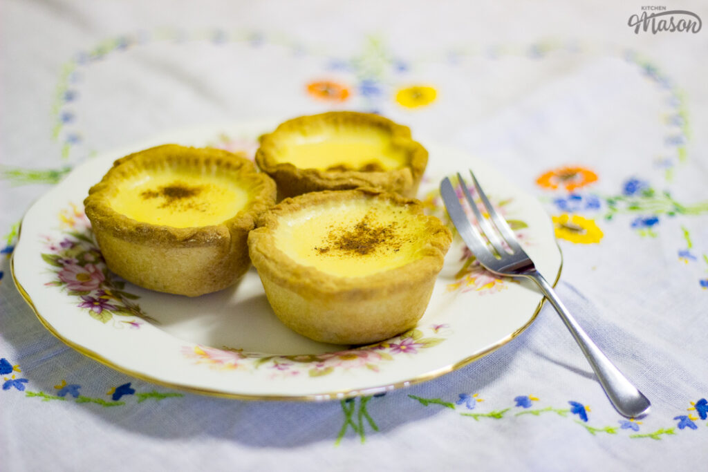 3 homemade egg custard tarts on a white floral plate with a dessert fork set over a white floral table cloth.