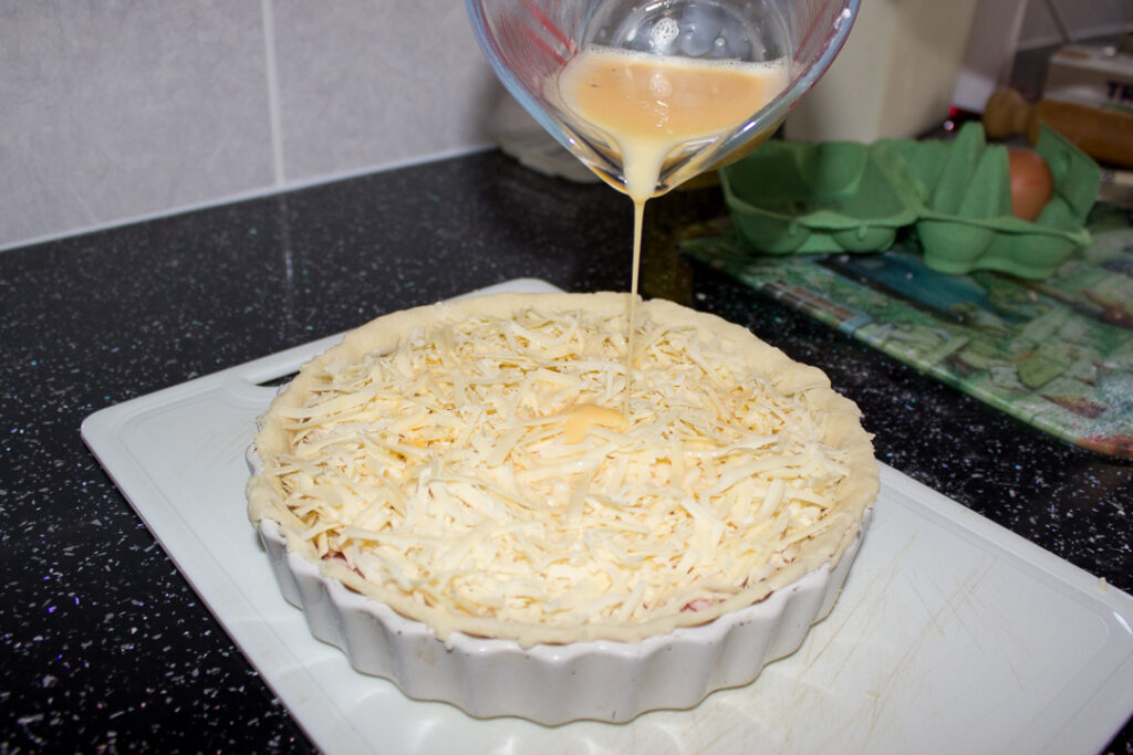 Seasoned egg and milk mixture being poured over the quiche on a kitchen worktop