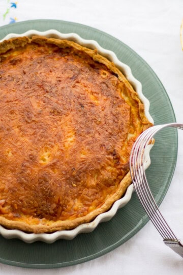Gran's ham and cheese quiche in a white baking dish on a green plate. Set on a white floral table cloth with a pastry blender, teacups and a saucer in the background.