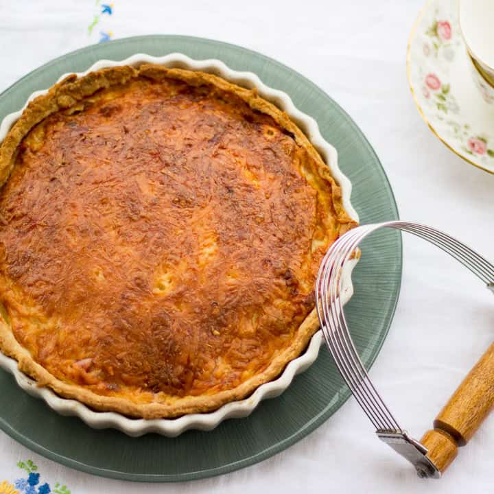 Gran's ham and cheese quiche in a white baking dish on a green plate. Set on a white floral table cloth with a pastry blender, teacups and a saucer in the background.