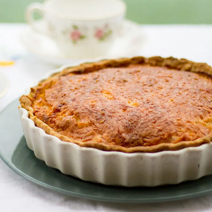 Gran's ham and cheese quiche in a white baking dish on a green plate. Set on a white floral table cloth with teacups and a saucer in the background.