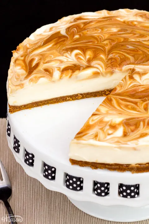 Close up of a Biscoff cheesecake with a slice out of it, set on a cake stand against a black backdrop.