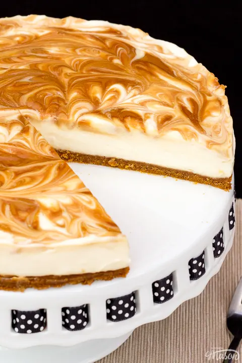 Close up of a Biscoff cheesecake with a slice out of it, set on a cake stand against a black backdrop.