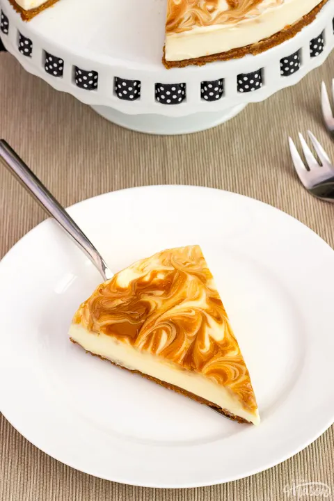 A slice of no bake Biscoff cheesecake on a white plate with a cake slice underneath. The rest of the cheesecake is on a cake stand in the background along with 2 forks.