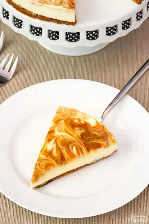 A slice of no bake Biscoff cheesecake on a white plate with a cake slice underneath. The rest of the cheesecake is on a cake stand in the background along with 2 forks.
