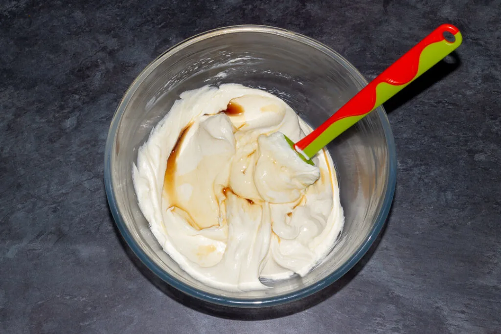 Cream cheese and caster sugar beaten together with vanilla in a glass bowl with a spatula on a kitchen worktop