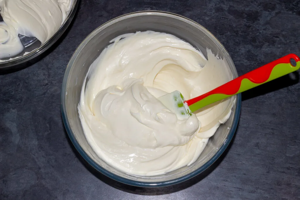 Whipped cream being folded into the cream cheese mixture in a glass bowl with a spatula on a kitchen worktop.